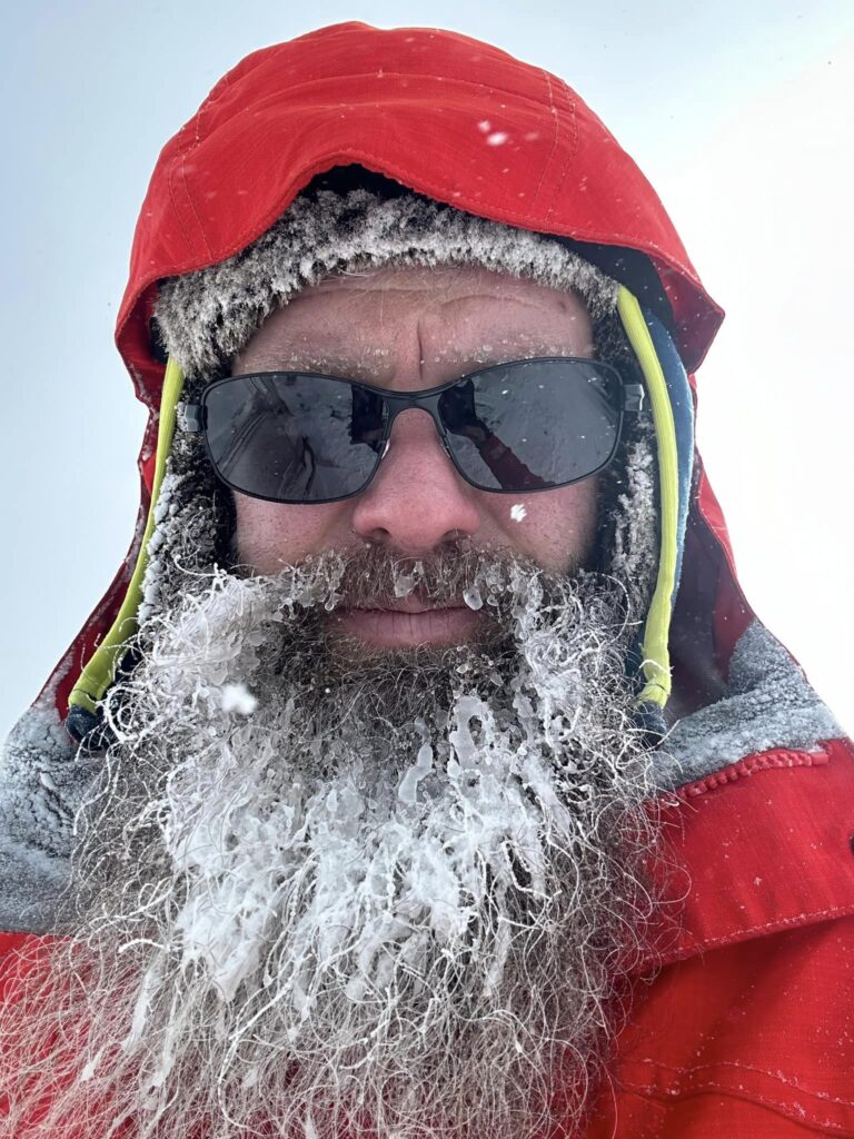 Photo shows a close up headshot of Graham Lau in the winter with an ice covered beard and sunglasses and winter wear.